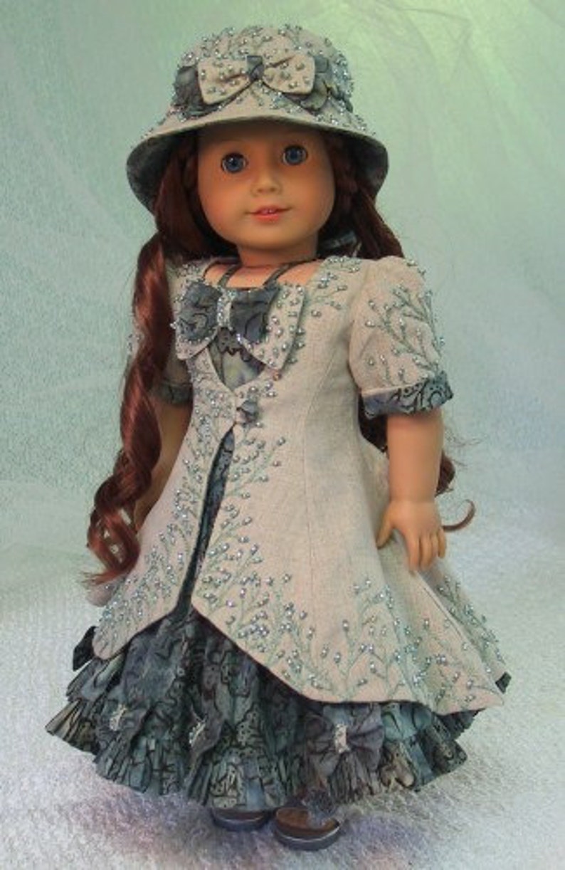 May include: A doll wearing a light gray dress with a floral pattern and a light blue hat with a bow. The doll has long red hair and blue eyes.
