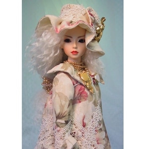 May include: A porcelain doll with long, white, curly hair, wearing a floral print coat and hat with lace trim. The doll has a pearl necklace and a gold-colored brooch. The doll's lips are painted red.