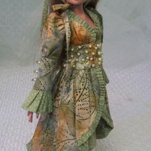 以下が含まれることがあります： 緑と茶色の模様のドレスを着た人形。お揃いのヘッドピースとパンツ付き。ドレスはパールで装飾され、フリル袖が付いています。人形は長い茶色の髪で、白い靴を履いています。