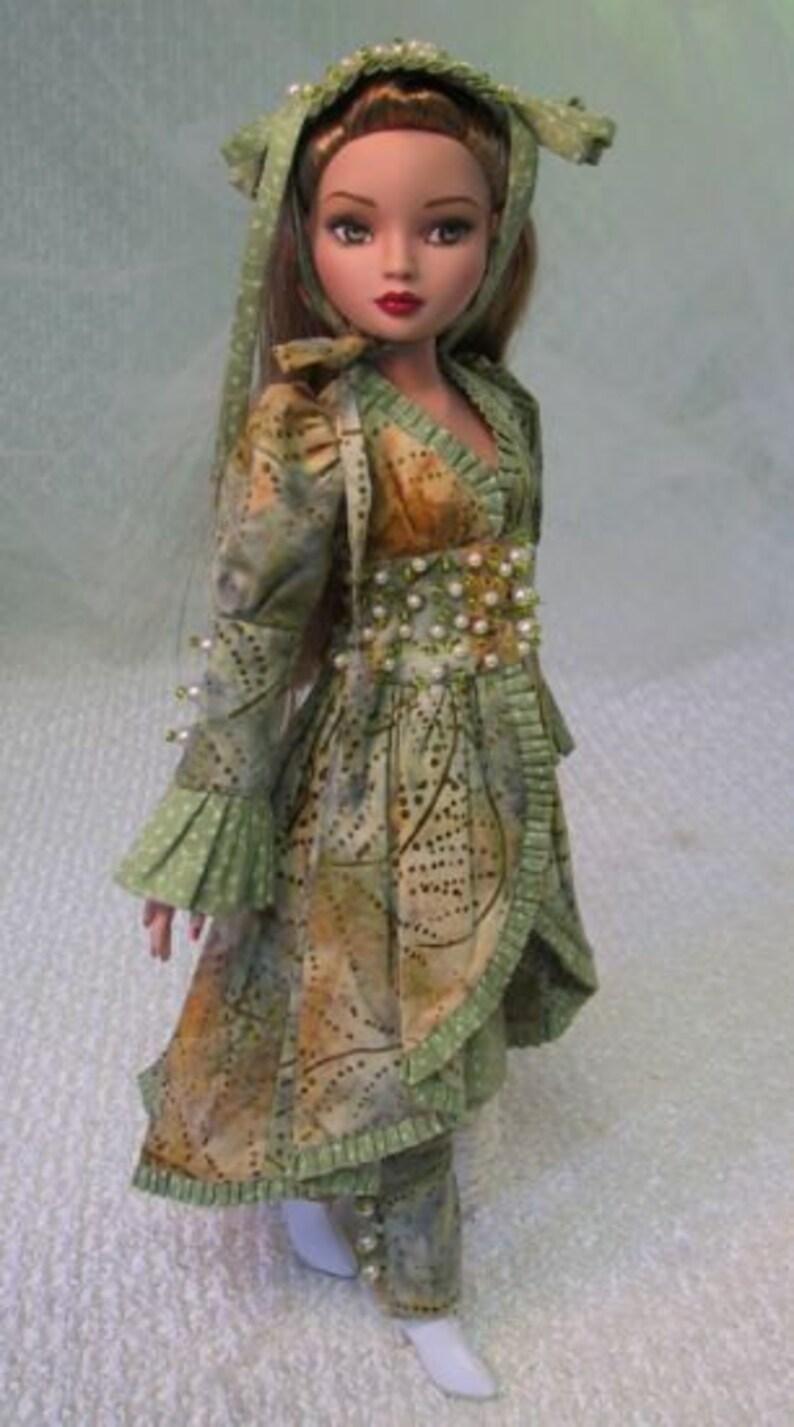 以下が含まれることがあります： 緑と茶色の模様のドレスを着た人形。お揃いのヘッドピースとパンツ付き。ドレスはパールで装飾され、フリル袖が付いています。人形は長い茶色の髪で、白い靴を履いています。