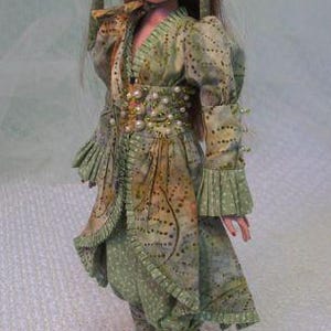 以下が含まれることがあります： 緑と茶色の模様の衣装を着た人形。お揃いのヘッドピース付き。衣装にはロングジャケット、パンツ、パールで飾られたベルトが含まれます。人形は白い靴を履いています。