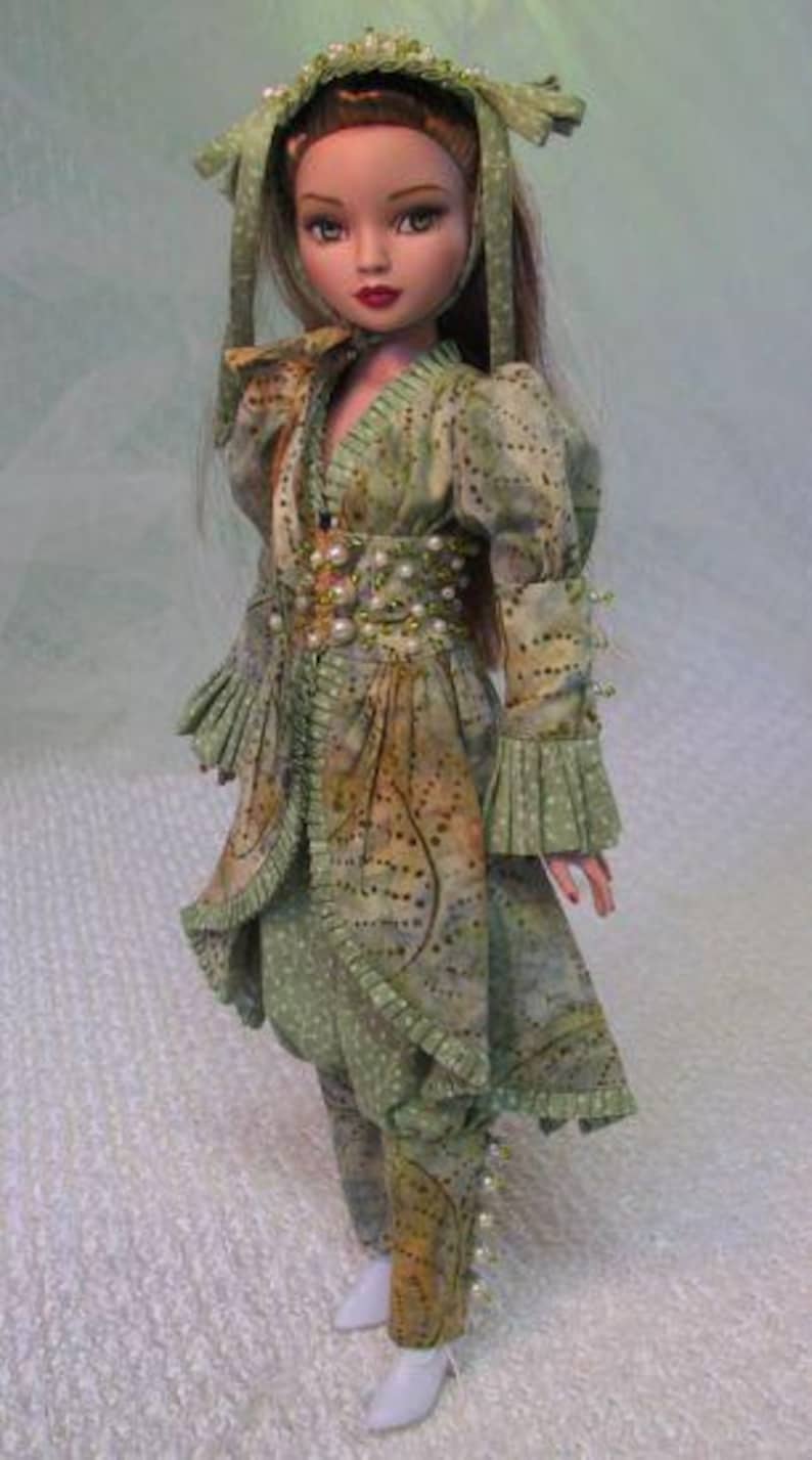 以下が含まれることがあります： 緑と茶色の模様の衣装を着た人形。お揃いのヘッドピース付き。衣装にはロングジャケット、パンツ、パールで飾られたベルトが含まれます。人形は白い靴を履いています。