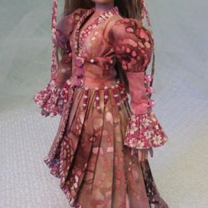 以下が含まれることがあります： ピンクと茶色の模様のドレスとヘッドピースを着た人形。ドレスはVネック、長袖、プリーツスカートです。人形は長い茶色の髪と白い靴を履いています。衣装はピンクのビーズで飾られています。全体的なデザインは花柄です。