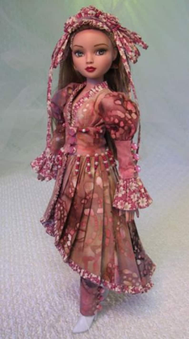 以下が含まれることがあります： ピンクと茶色の模様のドレスとヘッドピースを着た人形。ドレスはVネック、長袖、プリーツスカートです。人形は長い茶色の髪と白い靴を履いています。衣装はピンクのビーズで飾られています。全体的なデザインは花柄です。