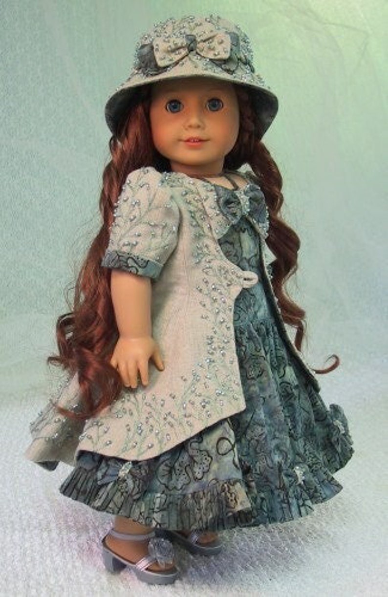 May include: A doll wearing a blue and white dress with a matching jacket and a light blue hat with a bow. The doll has long red hair and blue eyes.