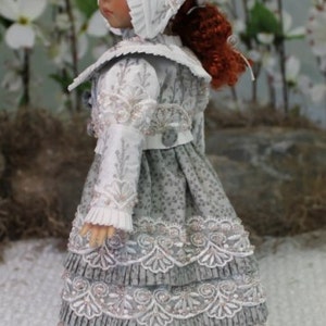 Puede incluir: Una mu&ntilde;eca de porcelana que lleva un vestido blanco y gris con ribetes de encaje y un gorro blanco. El vestido tiene una falda con volantes y el gorro tiene un ribete de encaje blanco.