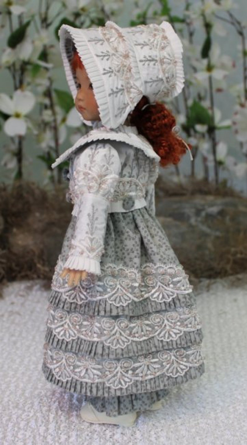 Puede incluir: Una mu&ntilde;eca de porcelana que lleva un vestido blanco y gris con ribetes de encaje y un gorro blanco. El vestido tiene una falda con volantes y el gorro tiene un ribete de encaje blanco.