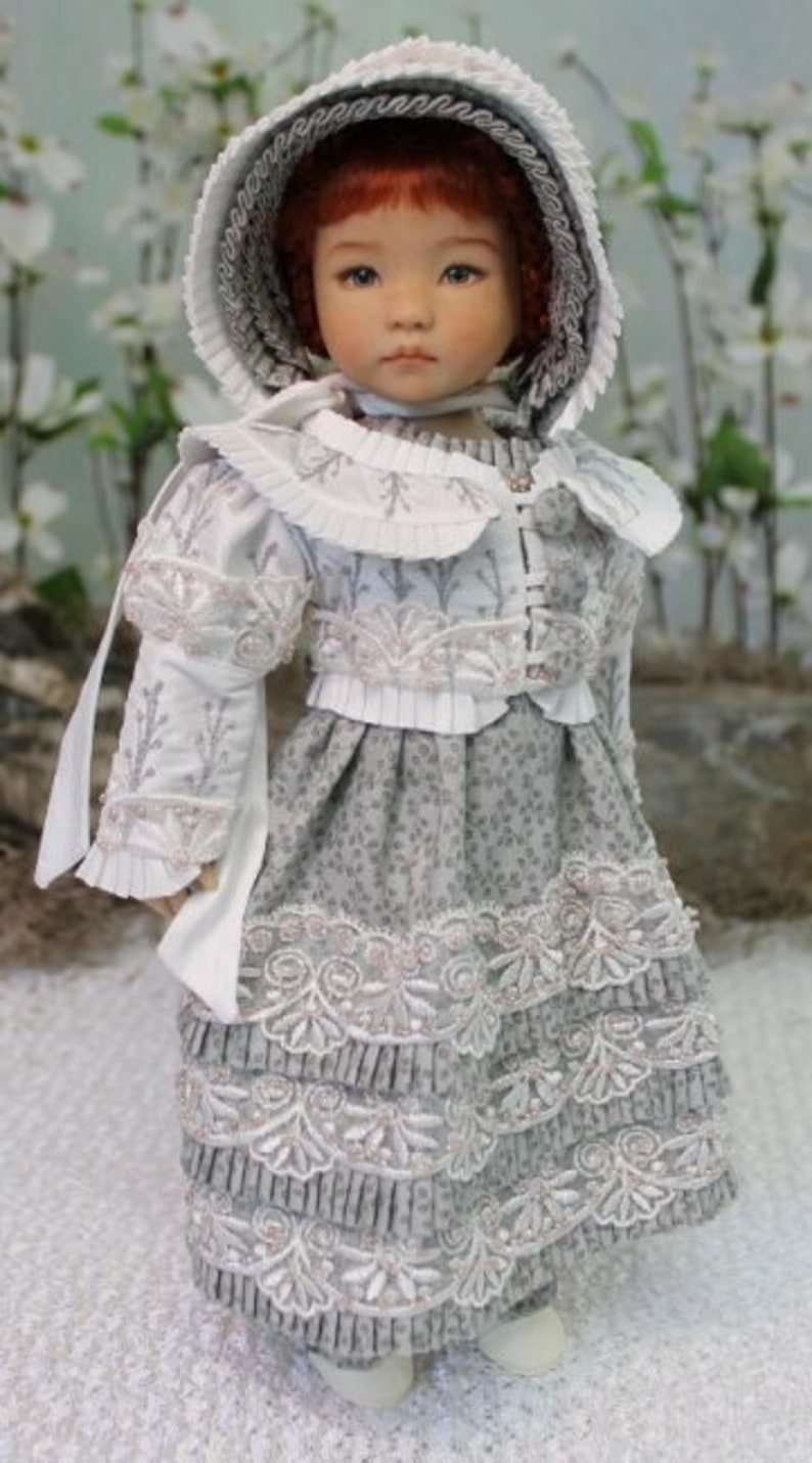 Puede incluir: Una mu&ntilde;eca de porcelana que lleva un vestido blanco y gris con ribetes de encaje y un gorro blanco. La mu&ntilde;eca tiene el pelo rojo y los ojos azules.