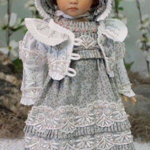 Puede incluir: Una mu&ntilde;eca de porcelana que lleva un vestido blanco y gris con ribetes de encaje y un gorro blanco. La mu&ntilde;eca tiene el pelo rojo y los ojos azules.