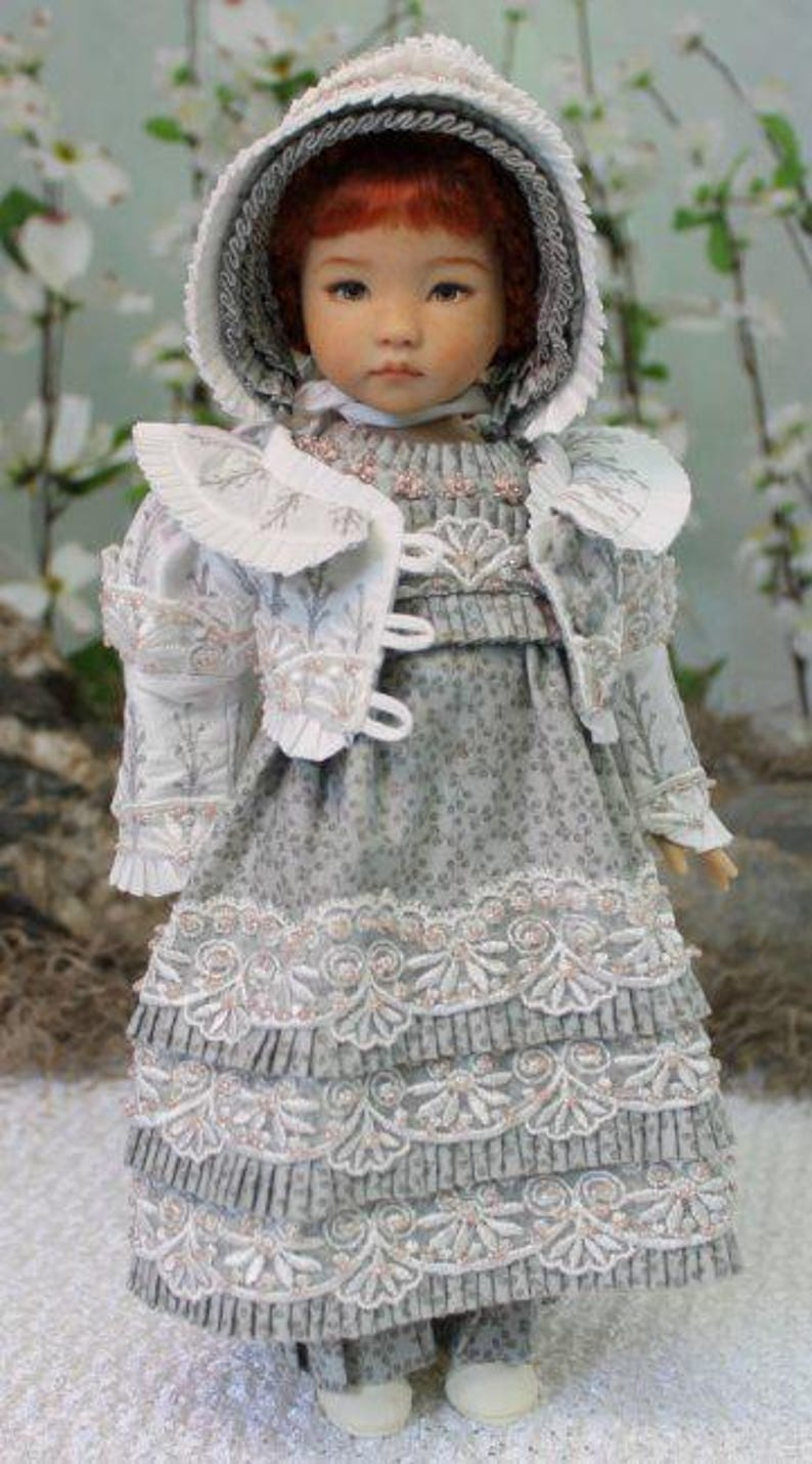 Puede incluir: Una mu&ntilde;eca de porcelana que lleva un vestido blanco y gris con ribetes de encaje y un gorro blanco. La mu&ntilde;eca tiene el pelo rojo y los ojos azules.