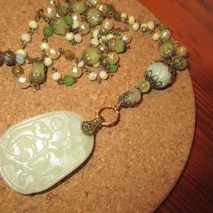 Puede incluir: Un collar largo de cuentas verdes y blancas con un colgante de jade tallado. El colgante presenta un dise&ntilde;o floral y est&aacute; unido al collar con una cadena dorada.