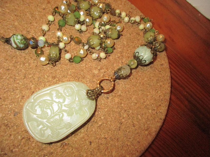 Puede incluir: Un collar largo de cuentas verdes y blancas con un colgante de jade tallado. El colgante presenta un dise&ntilde;o floral y est&aacute; unido al collar con una cadena dorada.