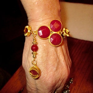 May include: A gold bracelet with a dangling charm features three large red gemstone cabochons. The bracelet is adorned with smaller gold beads and accents.