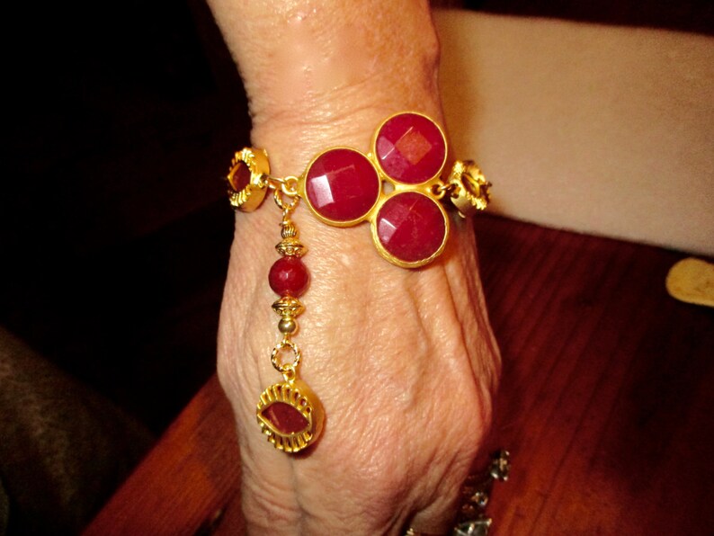 May include: A gold bracelet with a dangling charm features three large red gemstone cabochons. The bracelet is adorned with smaller gold beads and accents.