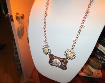 May include: A copper chain necklace with white pearl accents and a large, ornate pendant featuring a white, floral design.