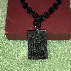 May include: A black chain necklace with a rectangular pendant featuring a gold-toned Buddha figure in a seated meditation pose. The pendant has intricate details and a black background.