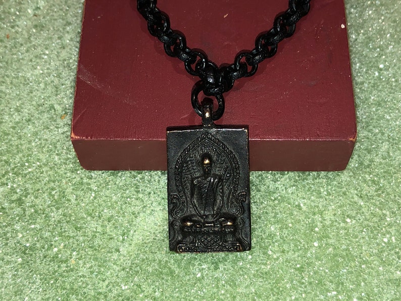 May include: A black chain necklace with a rectangular pendant featuring a gold-toned Buddha figure in a seated meditation pose. The pendant has intricate details and a black background.