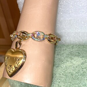 May include: A gold chain bracelet with a heart-shaped locket charm. The chain is adorned with colorful gemstones. The locket has a floral design.