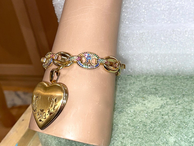 May include: A gold chain bracelet with a heart-shaped locket charm. The chain is adorned with colorful gemstones. The locket has a floral design.