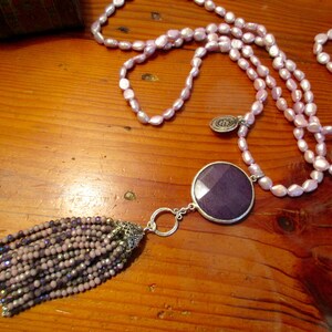 May include: A long necklace made of pink pearls with a large purple stone pendant and a beaded tassel. The necklace is on a wooden surface.
