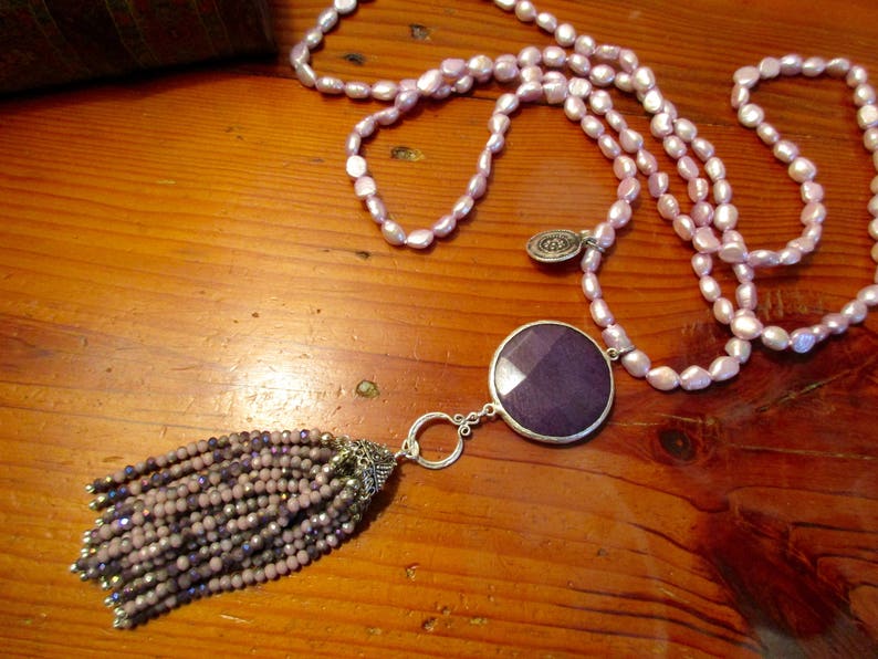 May include: A long necklace made of pink pearls with a large purple stone pendant and a beaded tassel. The necklace is on a wooden surface.