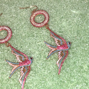 Puede incluir: Un par de pendientes de tono dorado con un dise&ntilde;o circular y un colgante de p&aacute;jaro golondrina. Los pendientes est&aacute;n adornados con cristales rosas y azules.