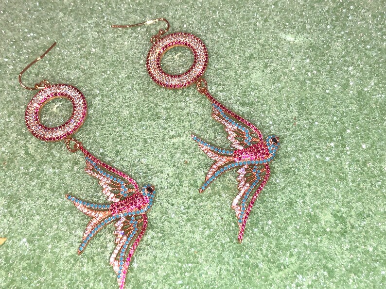 Puede incluir: Un par de pendientes de tono dorado con un dise&ntilde;o circular y un colgante de p&aacute;jaro golondrina. Los pendientes est&aacute;n adornados con cristales rosas y azules.