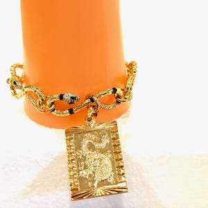 May include: A gold bracelet with a snake chain and a charm featuring a dragon design. The charm is a rectangular pendant with a detailed dragon image.