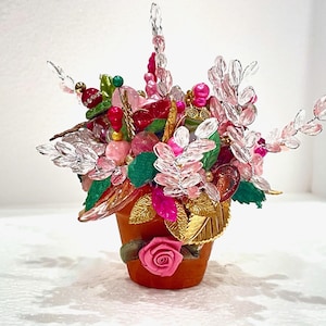 May include: A small terracotta pot with a pink rose on the front, filled with a colorful arrangement of pink, red, and green beads and crystals.