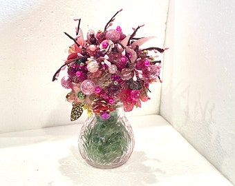 May include: A pink and white floral arrangement in a clear glass vase. The flowers are made of beads and other decorative elements.