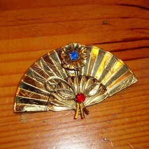 Puede incluir: Un broche de forma de abanico de color dorado con un adorno de pedrer&iacute;a azul y roja.