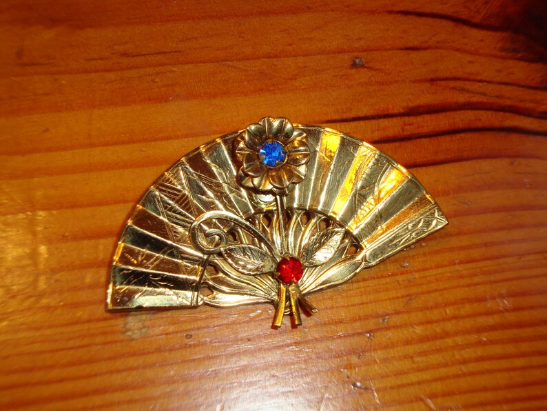 Puede incluir: Un broche de forma de abanico de color dorado con un adorno de pedrer&iacute;a azul y roja.