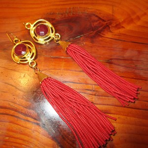 May include: A pair of gold earrings with red tassel drops. The earrings feature a round, gold setting with a red gemstone in the center.