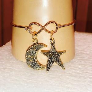 May include: Gold infinity bracelet with a crescent moon and star charm. The charms have a textured, swirling design.