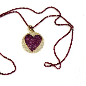 May include: A gold necklace with a round pendant featuring a heart-shaped design. The pendant is covered in small, sparkling red and clear stones. The necklace is made of small, red beads.
