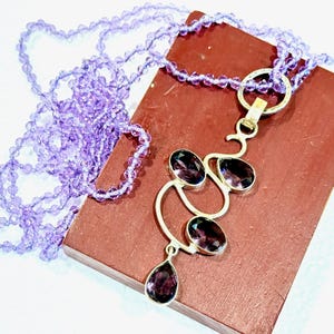 May include: A silver necklace with a pendant featuring a cluster of amethyst gemstones in a modern abstract design. The necklace is paired with a strand of light purple beads.