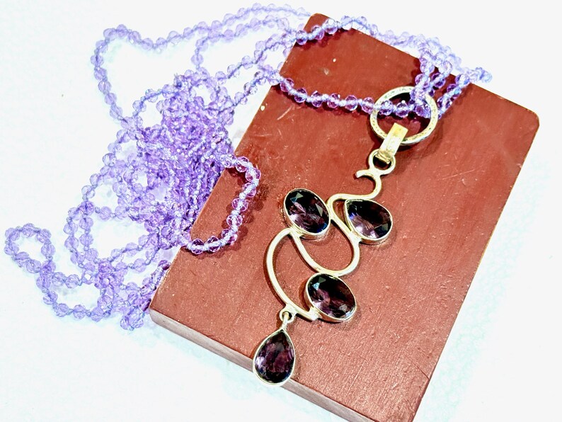 May include: A silver necklace with a pendant featuring a cluster of amethyst gemstones in a modern abstract design. The necklace is paired with a strand of light purple beads.