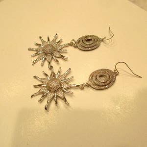 May include: A pair of silver earrings with a sunburst design and a circular design. The sunburst design is made of small, sparkling stones. The circular design is made of three concentric circles of stones.