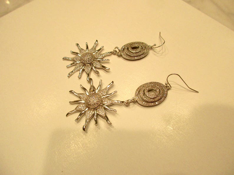 May include: A pair of silver earrings with a sunburst design and a circular design. The sunburst design is made of small, sparkling stones. The circular design is made of three concentric circles of stones.