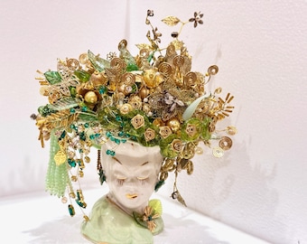 May include: A ceramic bust of a woman with closed eyes, wearing a green and gold floral crown made of wire, beads, and other decorative elements.