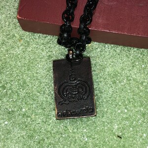 May include: A black chain necklace with a rectangular pendant. The pendant is made of dark metal and has a raised design of a symbol with the word "พระเครื่อง" written in Thai script.