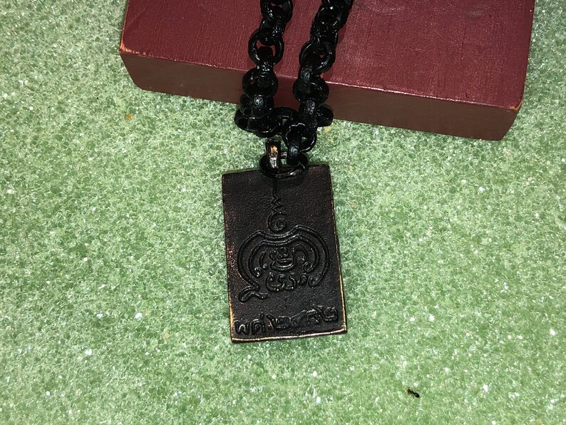 May include: A black chain necklace with a rectangular pendant. The pendant is made of dark metal and has a raised design of a symbol with the word "พระเครื่อง" written in Thai script.