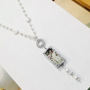 Pu&ograve; includere: Una collana di perle bianche con un ciondolo d'argento che presenta un'immagine incorniciata di una donna con un abito bianco. Il ciondolo &egrave; attaccato a un cerchio d'argento con piccole pietre trasparenti.