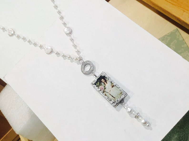 Pu&ograve; includere: Una collana di perle bianche con un ciondolo d'argento che presenta un'immagine incorniciata di una donna con un abito bianco. Il ciondolo &egrave; attaccato a un cerchio d'argento con piccole pietre trasparenti.