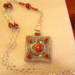 May include: A silver chain necklace with a square pendant featuring a red coral stone and intricate silver designs. The necklace has multiple strands of chain and red coral beads.