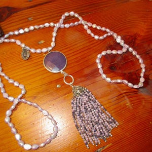 May include: A long, double-strand necklace made of light purple oval beads with a large, dark purple pendant and a tassel of smaller purple beads.