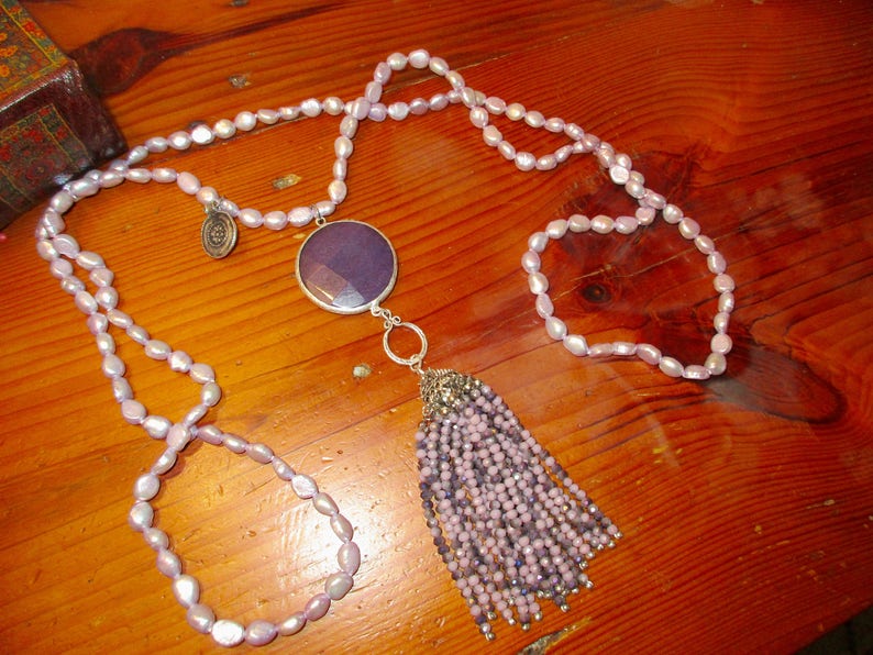 May include: A long, double-strand necklace made of light purple oval beads with a large, dark purple pendant and a tassel of smaller purple beads.
