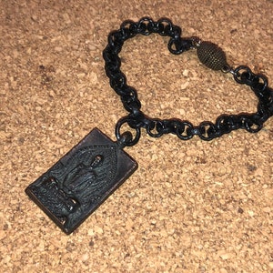 May include: A black chain bracelet with a rectangular pendant featuring a detailed image of a seated Buddha. The pendant is made of a dark metal and has a slightly aged look. The bracelet has a small, round, bronze-colored bead at the clasp.