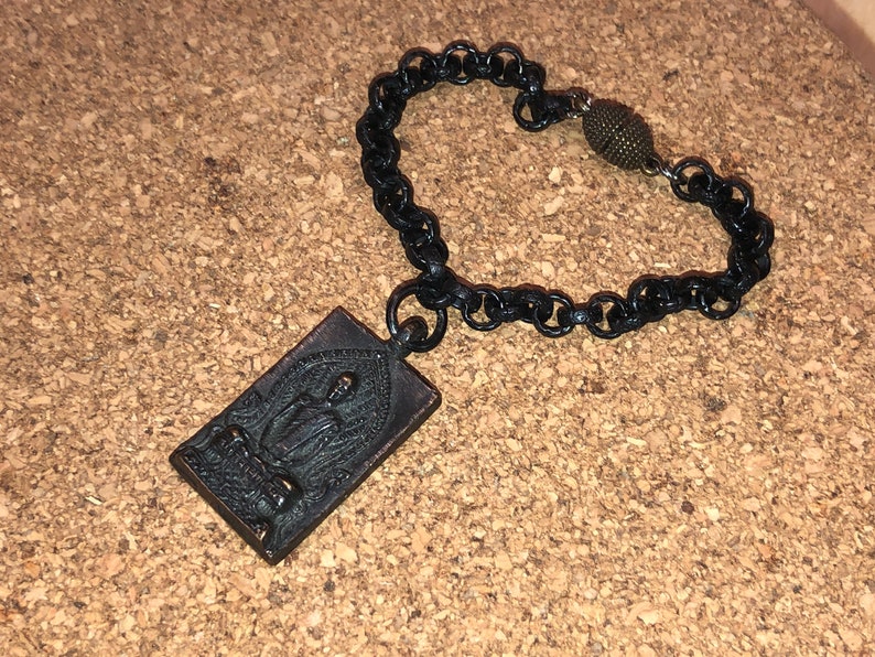 May include: A black chain bracelet with a rectangular pendant featuring a detailed image of a seated Buddha. The pendant is made of a dark metal and has a slightly aged look. The bracelet has a small, round, bronze-colored bead at the clasp.