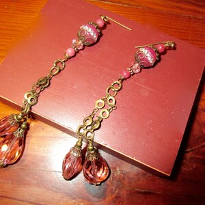 May include: A pair of long, pink and gold earrings with multiple beads and a dangling teardrop shape. The earrings are on a red wooden surface.